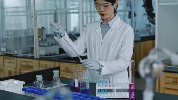 Ritratto medio di giovane donna cinese in camice bianco, occhiali e guanti aggiungendo gocce di reagente per testare il flaconcino con pipetta di vetro lunga, poi guardando la fotocamera e sorridendo - Filmati, video