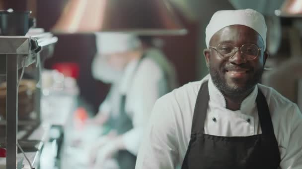 Portret radosnego czarnego kucharza w fartuchu i kapeluszu patrzącego w kamerę i szczęśliwie uśmiechającego się do pracy w kuchni restauracyjnej - Materiał filmowy, wideo