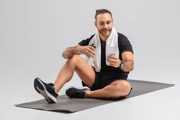Ένας άνδρας κάθεται σε ένα μαύρο στρώμα γυμναστικής, κρατώντας ένα μπουκάλι νερό, πιάνοντας την αναπνοή του μετά από μια συνεδρία προπόνηση. Φορώντας αθλητικά σορτς, αμάνικο μπλουζάκι και αθλητικά παπούτσια, χρησιμοποιώντας smartphone - Φωτογραφία, εικόνα