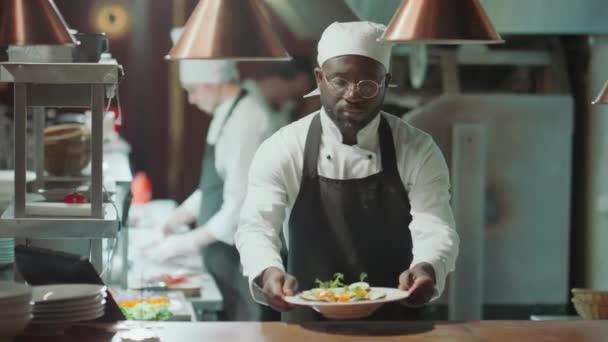 Portret młodego Afroamerykanina kucharza w fartuchu nakładanie talerza z serwowanym daniem na stół i pozowanie do aparatu z uśmiechem podczas pracy w kuchni restauracji - Materiał filmowy, wideo