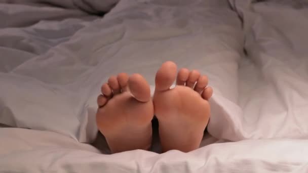 vrouwelijke benen film en dans tenen terwijl liggend in bed op een wit laken onder een deken. gelukkig vrouw meisje met blote benen voet is ontspannen en rusten op bed in een hotel slaapkamer in de avond - Video