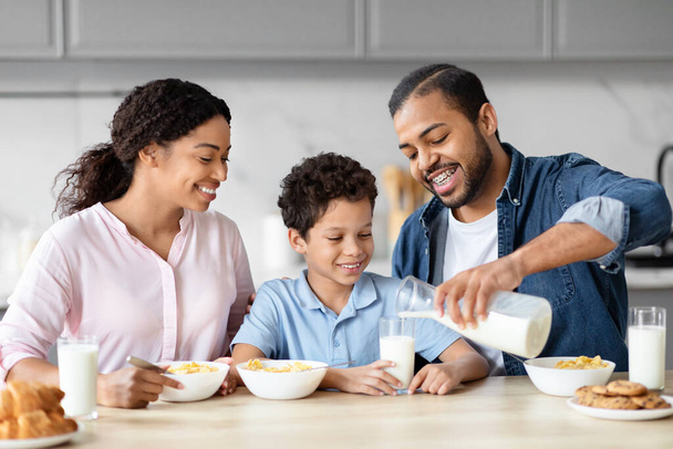 Eine afrikanisch-amerikanische Familie teilt einen hellen Moment, während der Vater Milch in ein Glas gießt, was die Wärme des Familienlebens in einer Küche verkörpert - Foto, Bild