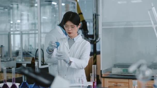 Medium shot van drukke Chinese vrouwelijke onderzoeker toe te voegen reagens met bol pipet om buis te testen, Afro-Amerikaanse mannelijke collega met tablet naderen en delen resultaat van experiment - Video