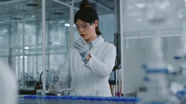 Genç Çinli kadın laboratuvar çalışanı, temiz çözeltiyle pembe kimyasal maddeyi şişeye boşaltıp dairesel hareketlerle karıştırırken, - Video, Çekim