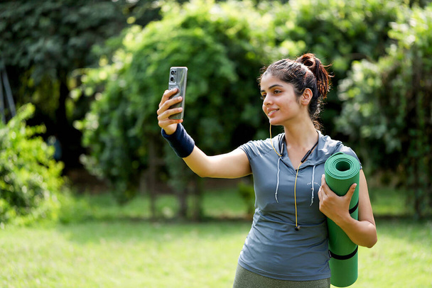 glimlachen sportschool outfit vrouw het nemen van selfie in park terwijl het houden van yoga mat in de hand - Foto, afbeelding