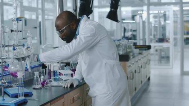 Mittlere Ganzseitenaufnahmen eines afroamerikanischen Chemikers, der im Labor arbeitet, neue synthetische Verbindungen entwickelt, mit Lösungen in Fläschchen experimentiert und Notizen auf einem digitalen Tablet macht - Filmmaterial, Video