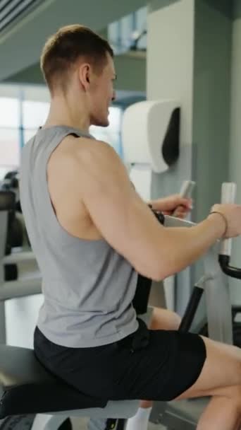 Young Man Focused on Building Muscle at a Modern Gym. Un jeune athlète masculin utilisant une machine à ramer au gymnase, s'est concentré sur ses objectifs de remise en forme, illustrant sa détermination et sa force. Vidéo verticale. - Séquence, vidéo