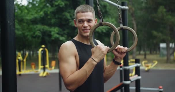 Portrait of young athletic man standing at sport ground and looking at camera. Handsome sporty guy prepare exercising with gymnastic rings. Muscle Up In Public Gym. Outdoors gym. - Footage, Video