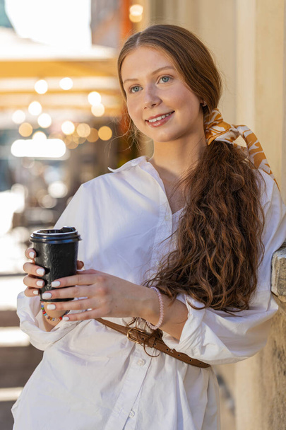 Happy redhead teenager girl enjoying morning coffee hot drink and smiling. Relaxing, taking a break. Young woman standing in urban city center street, drinking coffee to go. Town lifestyles. Vertical - Photo, Image