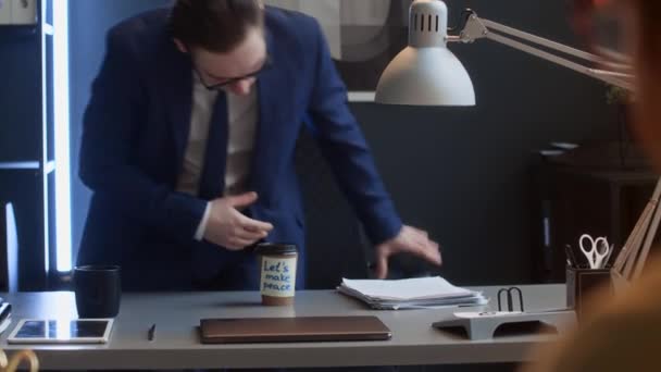 Colpo medio di dipendente aziendale maschio caucasico in giacca e cravatta che si avvicina alla scrivania con un mucchio di documenti, trova una tazza di caffè da asporto con nota pacifica, sorride, beve e mostra il pollice in alto - Filmati, video