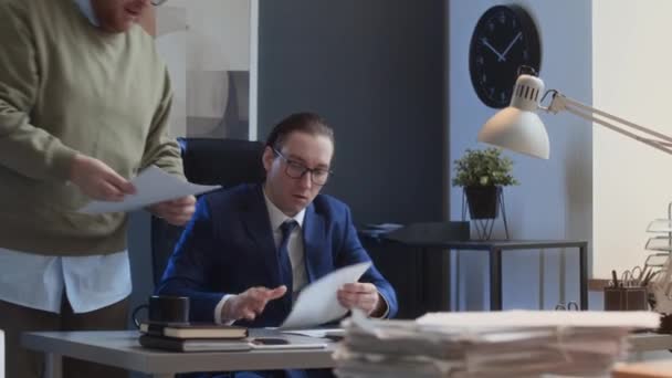 Medium shot of goofy Caucasian male employee walking up towards manager in suit sitting at desk, showing report draft, angry manager shouting, throwing pages in air and pushing him away - Footage, Video