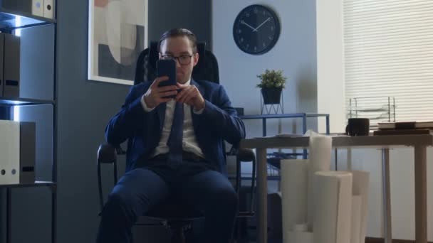 Μέτριο πλάνο του μέσου άντρα σταδιοδρομίας στο κοστούμι, γραβάτα και γυαλιά κάθεται στην καρέκλα στο γραφείο, χρησιμοποιώντας smartphone, στη συνέχεια να γλιστρήσει μέχρι συνάδελφο περπατώντας με στοίβα από χαρτιά και γέλιο βίαια - Πλάνα, βίντεο