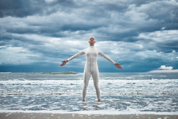 Щаслива дівчина без волосся з алопецією в білому футуристичному костюмі стоїть з розкиданими руками на пляжі, купатися океанськими хвилями, виступ лисої сильної жінки-художниці символізує самоприйняття - Фото, зображення