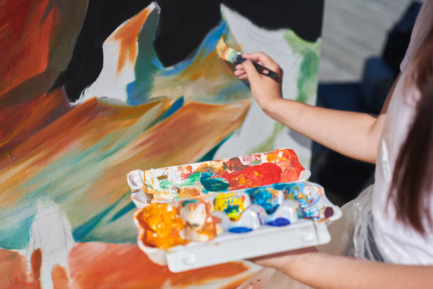 Γυναικείος ζωγράφος χέρι ζωγραφίζει με πάθος την εικόνα με πινέλο για υπαίθρια έκθεση δρόμου χρησιμοποιώντας ζωντανά χρώματα, από κοντά - Φωτογραφία, εικόνα