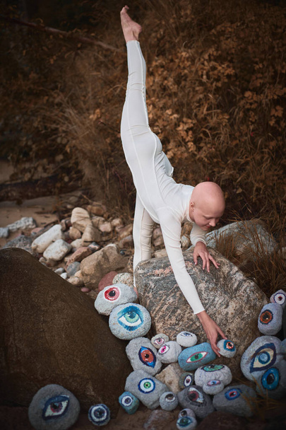 Flexible hairless girl with alopecia in white futuristic costume put her foot up and reaches hand for surreal landscape with lot of rocks with eyes, profound symbolism of embracing individuality - Photo, Image
