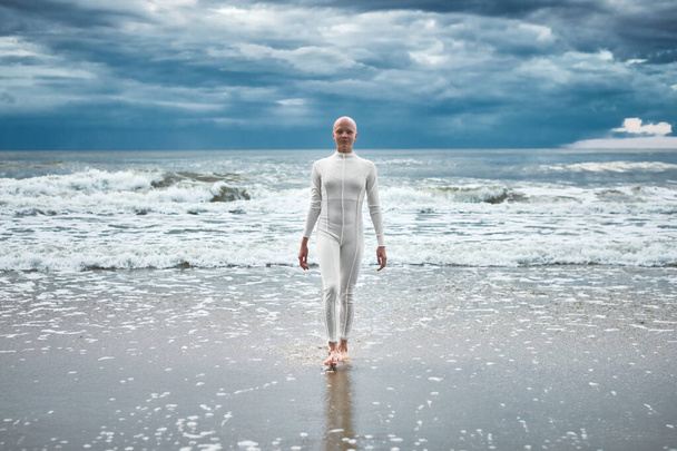 Bátor szőrtelen lány alopéciával fehér futurisztikus öltönyben kijön a hideg tengerből a homokos parton, metaforikus előadást ad a kopasz női művésznőnek az élet kihívásairól és az önelfogadásról. - Fotó, kép