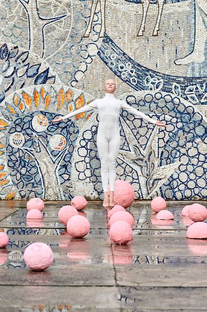 Young hairless girl ballerina with alopecia in white futuristic suit dancing outdoor and jumps among pink spheres on abstract mosaic Soviet background, symbolizes self expression - Photo, Image