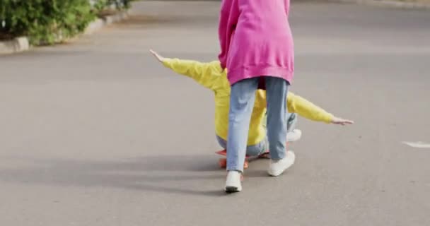 Ragazza spinge amico adolescente seduto su skateboard a cavalcare in avanti. Giovani migliori amiche si divertono nel parco urbano durante i fine settimana comuni - Filmati, video