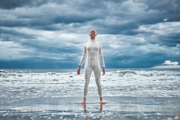 Felice ragazza senza peli con alopecia in abito bianco futuristico si erge sulla spiaggia bagnata dalle onde dell'oceano, performance metaforica di calva forte artista femminile, superare le sfide della vita e fiducia in se stessi - Foto, immagini
