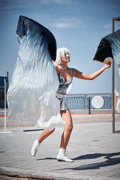 Młoda seksowna dziewczyna w przestrzeni srebrny mikro spódnica taniec z wentylatorem wydajność machając wdziękiem, kobieta taniec na świeżym powietrzu na nadmorskiej promenadzie tworząc spektakl zewnętrzny harmonizuje z rytmem fal morskich - Zdjęcie, obraz
