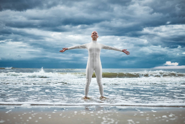 Glückliches haarloses Mädchen mit Alopezie im weißen futuristischen Anzug steht mit ausgebreiteten Armen am Strand, der von Meereswellen gebadet wird, Auftritt einer glatzköpfigen, starken Künstlerin, die die Selbstakzeptanz symbolisiert - Foto, Bild