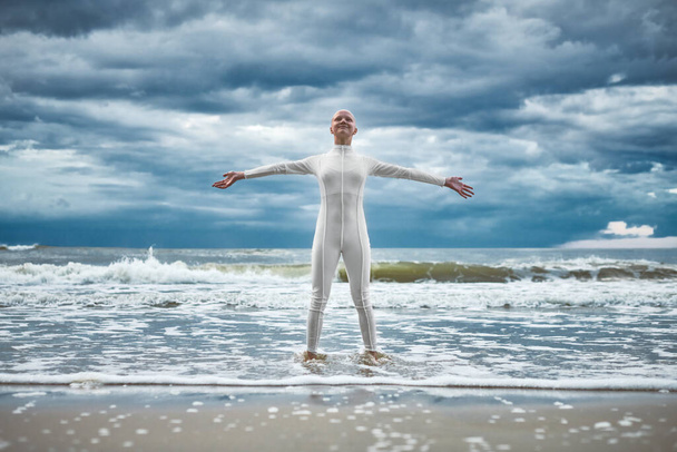 Glückliches haarloses Mädchen mit Alopezie im weißen futuristischen Anzug steht mit ausgebreiteten Armen am Strand, der von Meereswellen gebadet wird, Auftritt einer glatzköpfigen, starken Künstlerin, die die Selbstakzeptanz symbolisiert - Foto, Bild