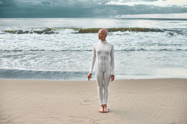 Ganztägiges Porträt eines jungen haarlosen Mädchens mit Alopezie im weißen futuristischen Anzug, das am Meeresstrand steht, metaphorische surreale Szene mit einem glatzköpfigen hübschen Teenager-Mädchen strahlt Zuversicht und einzigartige Schönheit aus - Foto, Bild