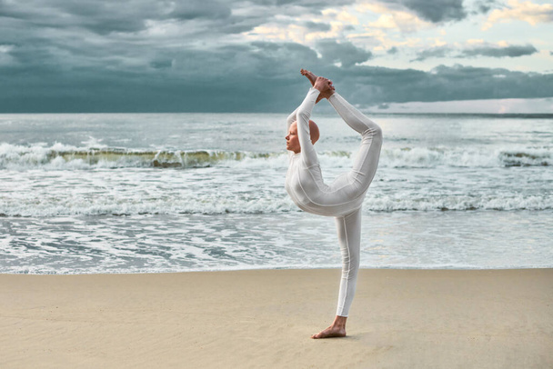 Giovane ballerina glabra con alopecia in tuta bianca futuristica che balla sulla spiaggia di sabbia, metaforica performance surreale con calva bella adolescente trasuda fiducia e bellezza unica - Foto, immagini