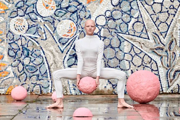 Девушка без волос с алопецией в белом футуристическом костюме танцует на открытом воздухе плавно держа розовый мяч на абстрактном мозаичном советском фоне, символизирует самовыражение и культурную самобытность - Фото, изображение