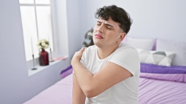 Молодий чоловік з дискомфортом потирає плечовий біль в інтер'єрі біло-фіолетової спальні. - Кадри, відео