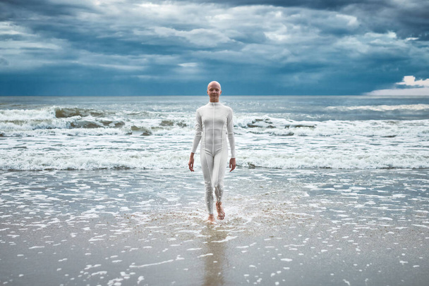 Tapferes haarloses Mädchen mit Alopezie im weißen futuristischen Anzug kommt aus dem kalten Meer am Sandstrand, metaphorische Performance der glatzköpfigen Künstlerin über die Bewältigung der Herausforderungen des Lebens und die Selbstakzeptanz - Foto, Bild