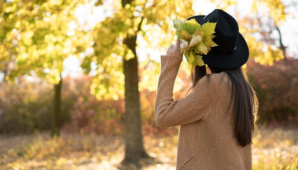 Женщина в черной шляпе и вязаном свитере ходит по осеннему парку с жёлтыми листьями в руках. Свобода и беззаботная концепция. Копирование пространства - Фото, изображение