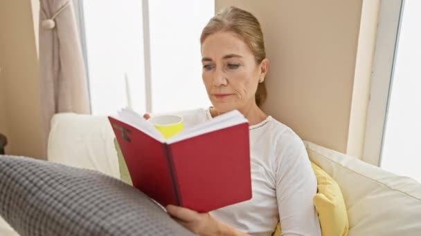 Mature woman reading book and drinking coffee in a cozy bedroom setting - Footage, Video
