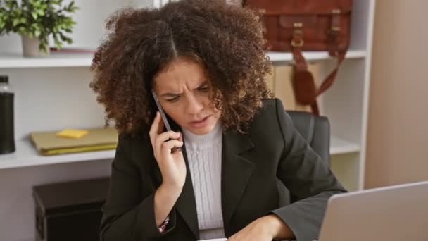 Een jonge Spaanse vrouw met krullend haar praat aan de telefoon in een kantoor setting, op zoek naar bezorgd. - Video