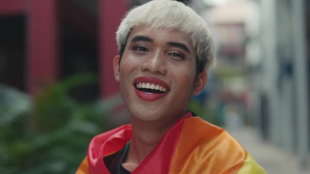 LGBT topluluğunun gülümseyen temsilcisi, Gökkuşağı Bayrağına Sarılı, Kamera İçin Duruşlar ve Pozisyonlar. Havalı saç stili ve Gökkuşağı Bayrağı ile dışarıda makyaj yapan mutlu adam. Çeşitlilik LGBT Kavramı - Video, Çekim