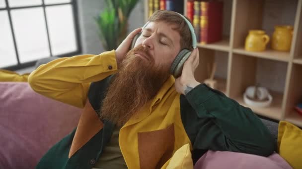 Расслабленный бородатый мужчина наслаждается музыкой в наушниках, сидя на красочном диване - Кадры, видео