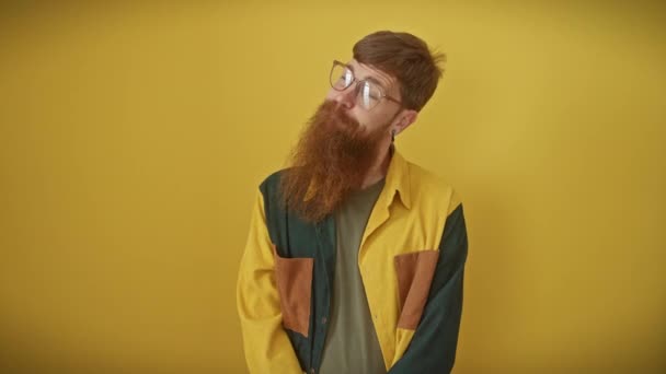 Cheerful young redhead guy, stylishly rocking glasses and shirt, caught in a smart thought while confidently standing and looking away on an isolated yellow background - Footage, Video