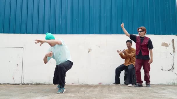 Fiatal utcai táncos hip-hop stílusban táncol multikulturális barátaival a falnál. Vonzó jóképű férfi nyújtja karjait az utcán, kék háttérrel. Szabadtéri sport 2024. hiphop. - Felvétel, videó