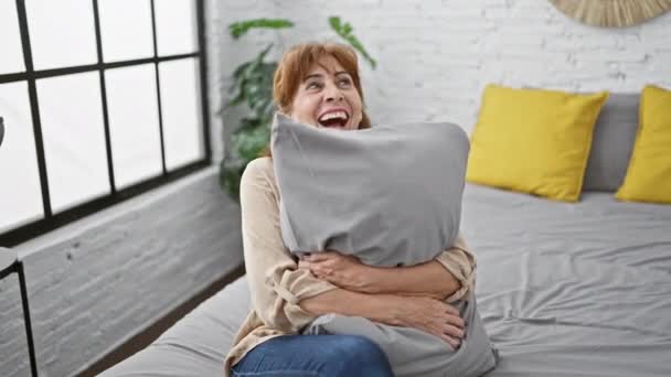 Femme d'âge moyen étreignant l'oreiller dans son lit, riant à haute voix à une blague drôle, rayonnant de joie dans sa chambre - Séquence, vidéo