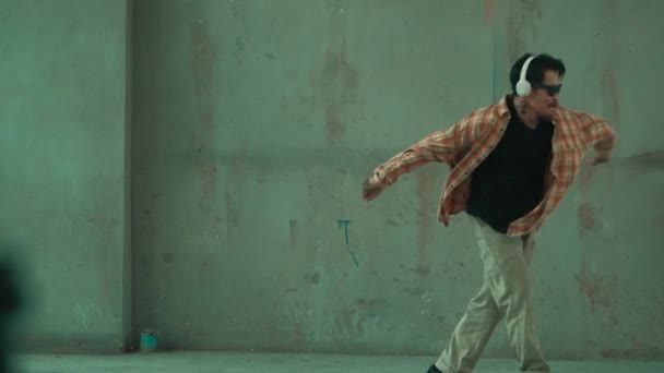 Aantrekkelijke Latijns-Amerikaanse danser in casual look praktijk straat dans, terwijl muziek luisteren vanaf de hoofdtelefoon op cement achtergrond. Motion shot van breakdancevoorstelling. Outdoor sport 2024. hiphop. - Video