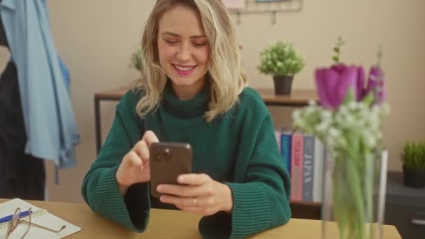 Usmívající se blondýnka používající svůj smartphone doma, obklopená květinami a brýlemi na dřevěném stole. - Záběry, video