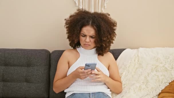 Hispanische junge Frau mit lockigem Haar benutzt Smartphone im Wohnzimmer der Wohnung, drückt Verwirrung aus, während sie auf der Couch sitzt. - Filmmaterial, Video