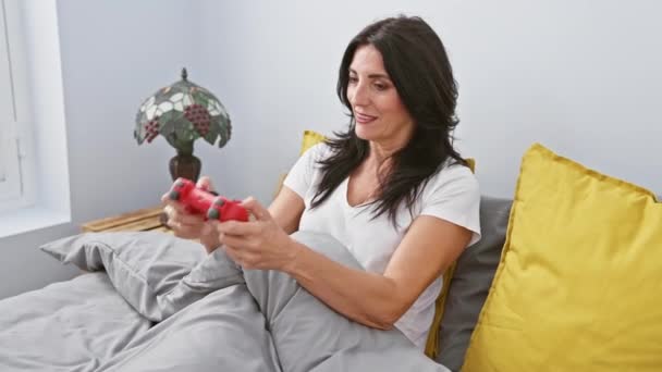 Ώριμη γυναίκα παίζει ένα βιντεοπαιχνίδι με χαρά και συγκέντρωση στο άνετο υπνοδωμάτιο της, που ενσωματώνει χαλάρωση και σύγχρονη ψυχαγωγία στο σπίτι. - Πλάνα, βίντεο