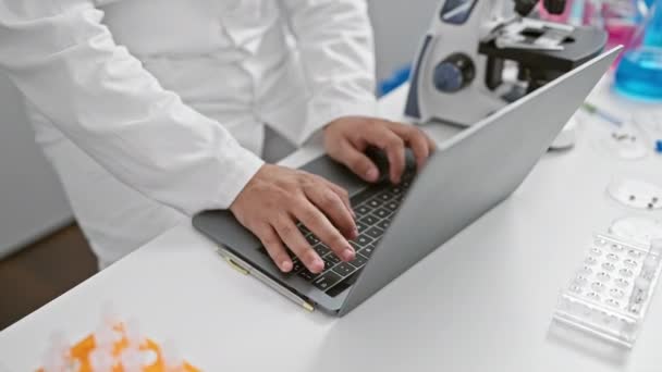 Een jongeman in een labjas werkt ijverig op een laptop in een modern laboratorium, met wetenschappelijke apparatuur om zich heen. - Video