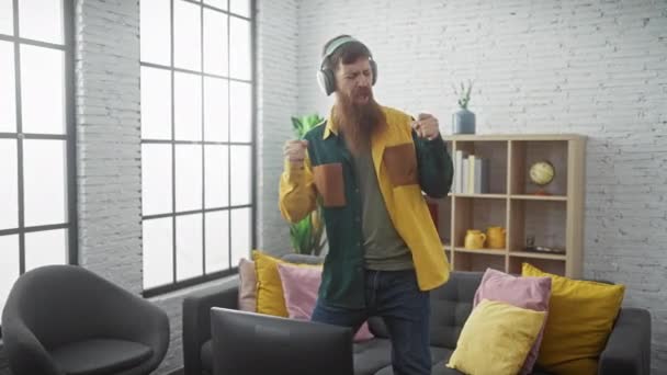Bearded young man in casual attire playfully dances in a modern interior while wearing headphones. - Footage, Video
