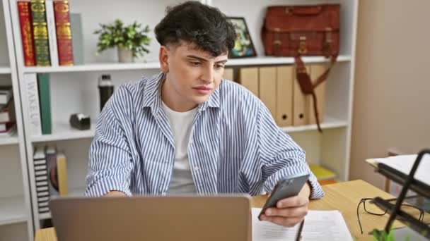 Νεαρός άντρας στέλνει μηνύματα σε ένα μοντέρνο γραφείο με λάπτοπ, χαρτιά και βιβλιοθήκη. - Πλάνα, βίντεο