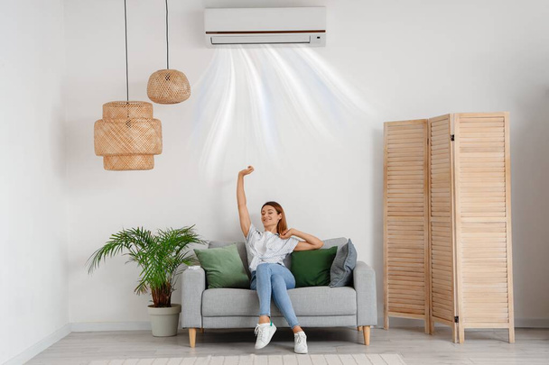 Junge Frau mit funktionierender Klimaanlage sitzt zu Hause auf Sofa - Foto, Bild