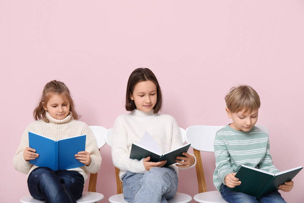Little children reading books while sitting on chairs against pink background - Foto, Imagen