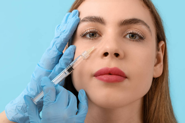 mooie jonge vrouw het ontvangen van filler injectie in gezicht op blauwe achtergrond - Foto, afbeelding