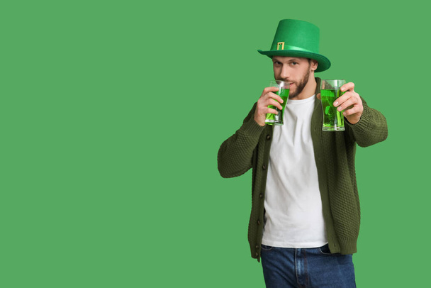 Молодой человек в шляпе лепрекона с бокалами пива на зеленом фоне. Празднование Дня Святого Патрика - Фото, изображение